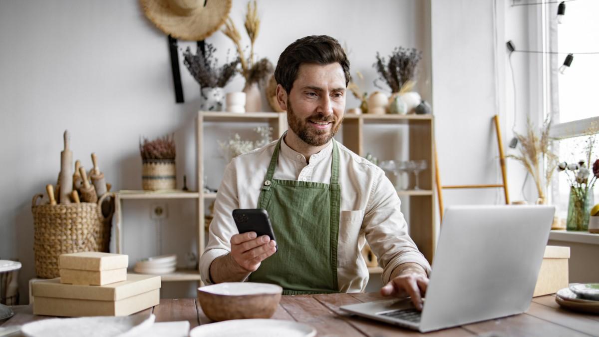 A young bearded male in a white shirt and green apron sits at his desk, holding his phone and typing on his laptop as he looks into the best payment systems for small businesses
